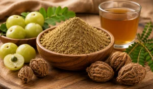 A wooden bowl filled with fine Triphala Churna powder surrounded by fresh green amla (Indian gooseberries), dried haritaki and bibhitaki fruits, and a glass of herbal triphala tea.